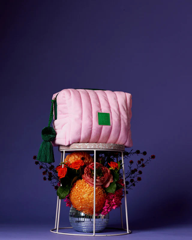 Pink quilted bag with a green tag on a stand with flowers against a purple background