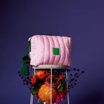 Pink quilted bag with a green tag on a stand with flowers against a purple background