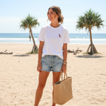 Woman on a beach holding a woven bag, with palm trees and ocean in the background