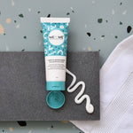 A squeeze tube of ME+ME Gentle Moisturiser moisturizer with its cap removed, placed on a dark gray textured mat with a squiggle of white cream beside it, against a speckled light blue background with a white towel on the right.