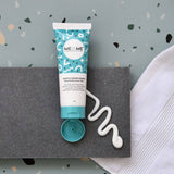 A squeeze tube of ME+ME Gentle Moisturiser moisturizer with its cap removed, placed on a dark gray textured mat with a squiggle of white cream beside it, against a speckled light blue background with a white towel on the right.