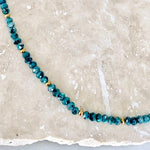 A beaded necklace made of faceted teal-blue beads separated by small gold spacer beads, displayed on a light beige stone surface.