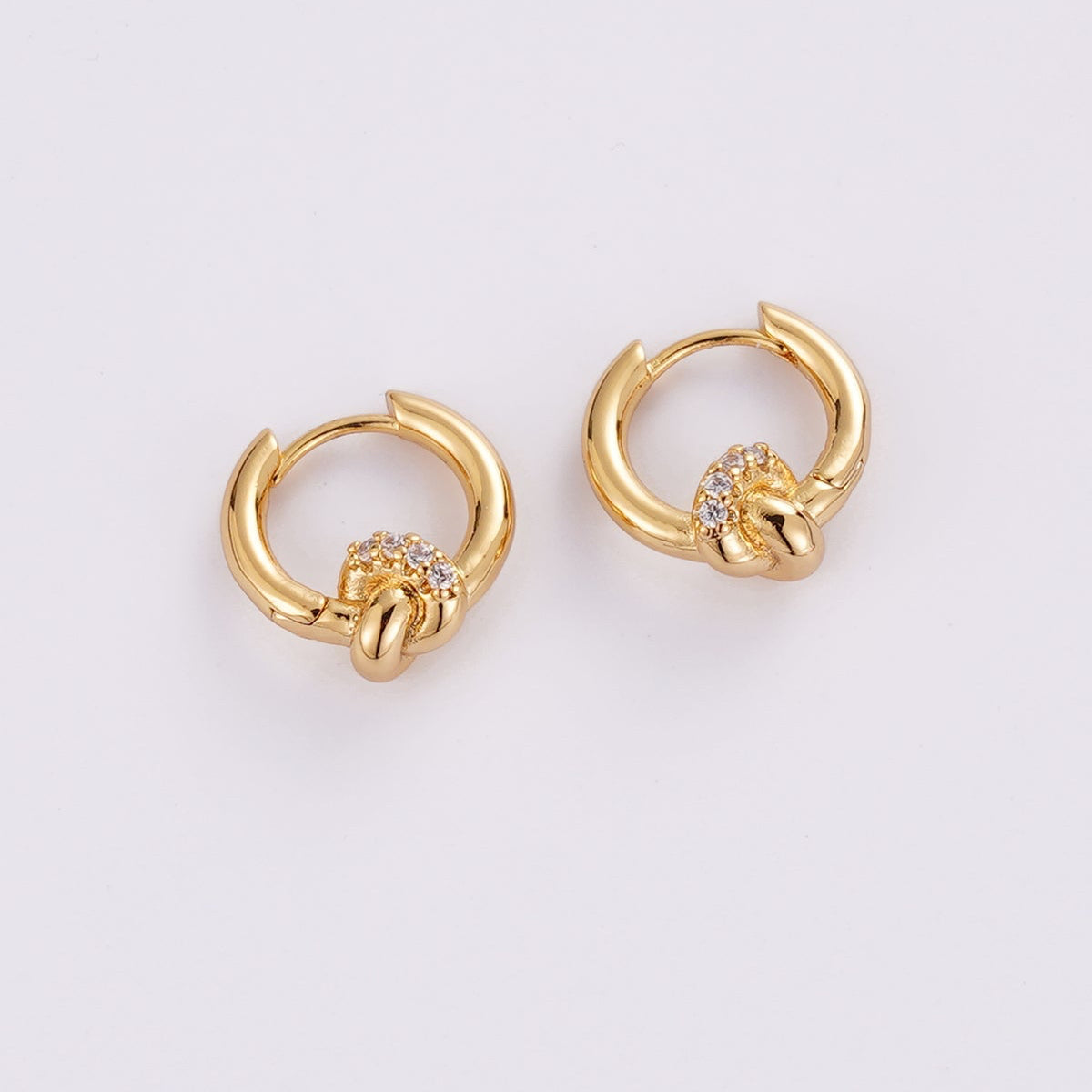 A pair of gold huggie hoop earrings with a knotted design and a small cluster of clear stones at the center, shown on a plain light gray background.