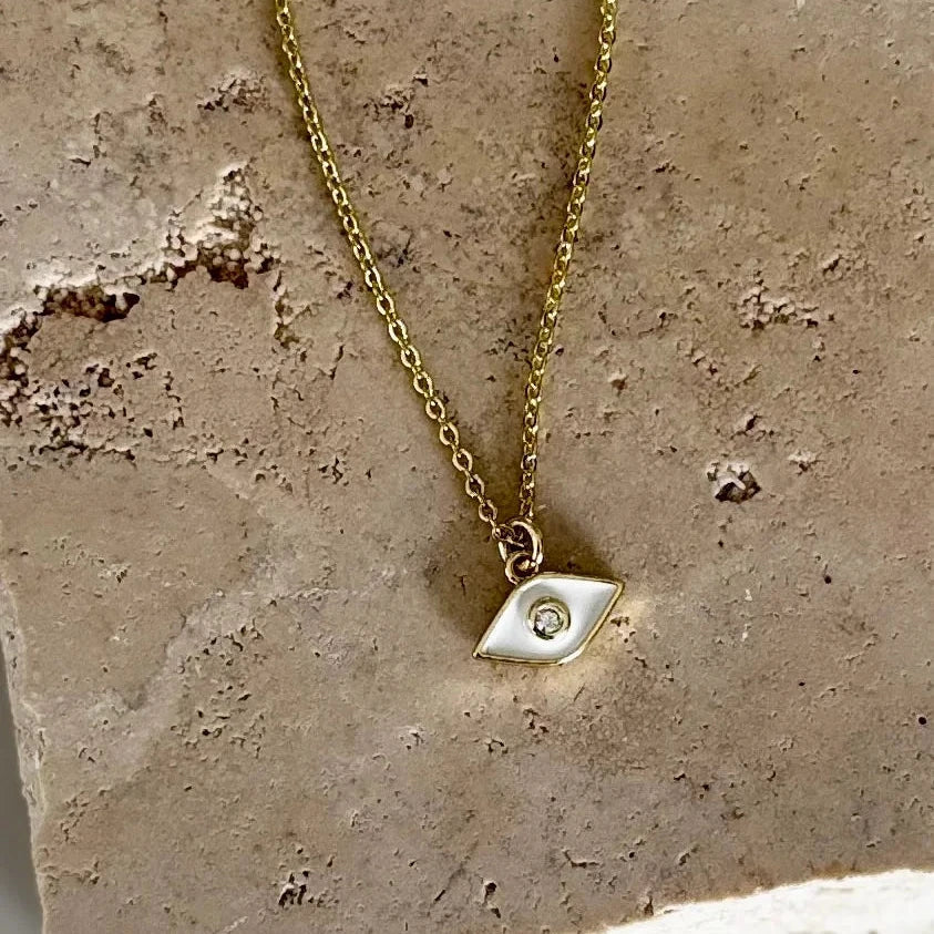 A delicate gold chain necklace with a small geometric silver pendant featuring a tiny clear stone, displayed on a textured beige stone slab.