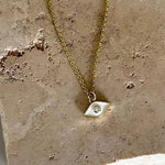 A delicate gold chain necklace with a small geometric silver pendant featuring a tiny clear stone, displayed on a textured beige stone slab.