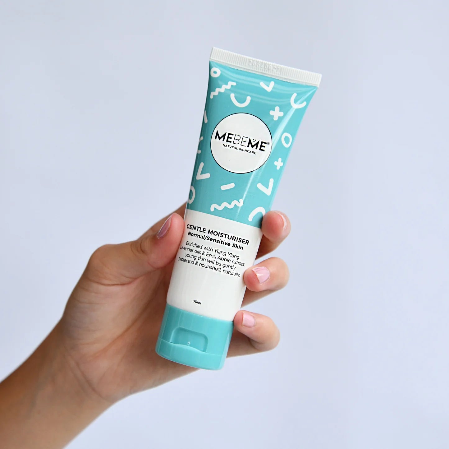 A hand holding a squeeze tube of MEBE ME Gentle Moisturiser against a light blue background.