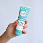 A hand holding a squeeze tube of MEBE ME Gentle Moisturiser against a light blue background.