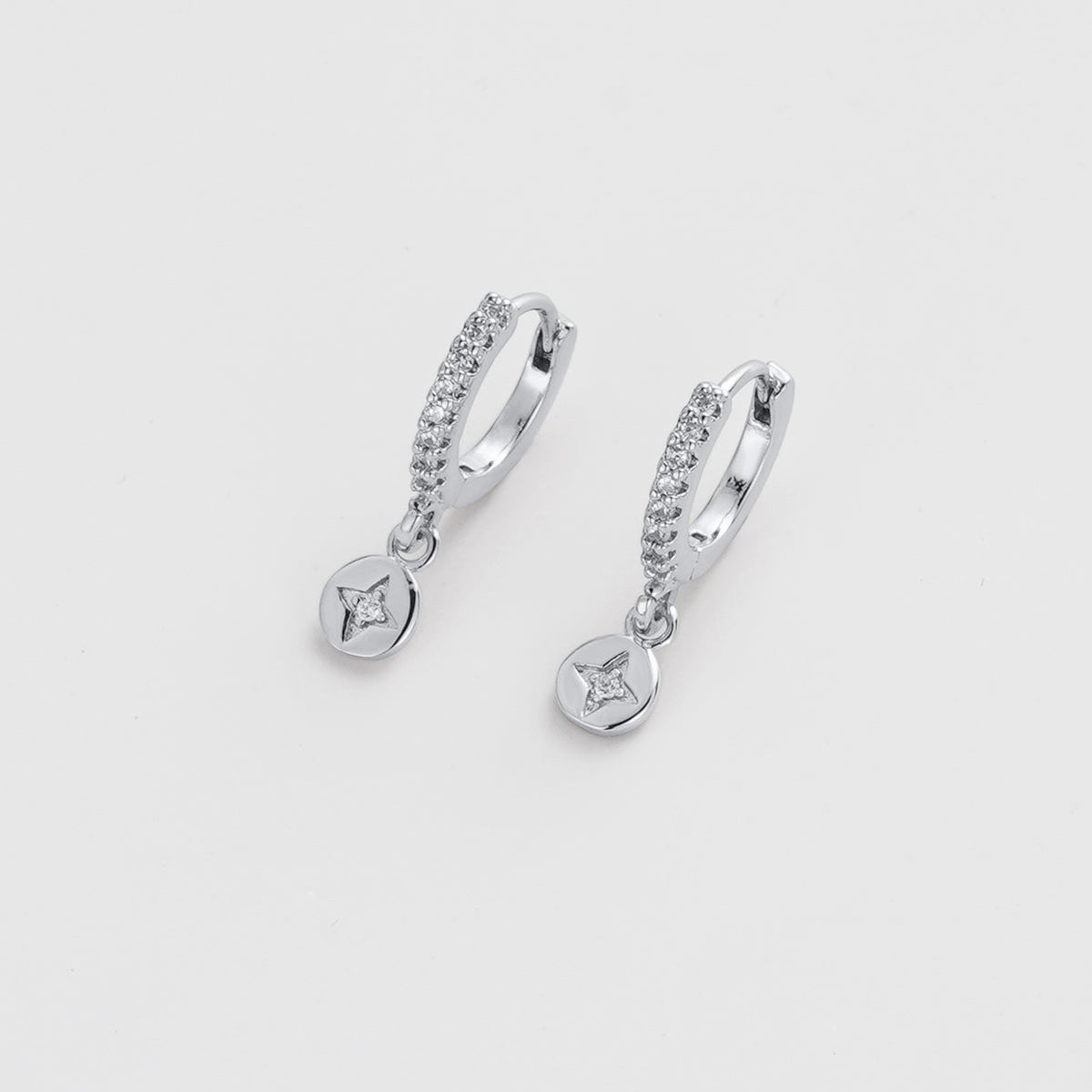 A pair of silver huggie hoop earrings with small clear stones and dangling round charms, shown on a light gray background.