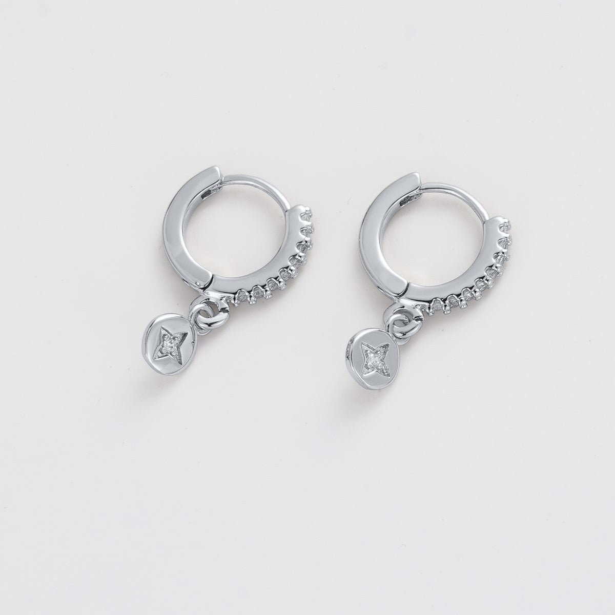A pair of silver huggie hoop earrings with small dangling charms, photographed on a clean, light gray background.