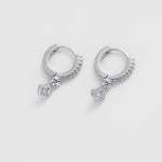 A pair of silver huggie hoop earrings with small dangling charms, photographed on a clean, light gray background.