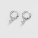A pair of silver huggie hoop earrings with small dangling charms, photographed on a clean, light gray background.