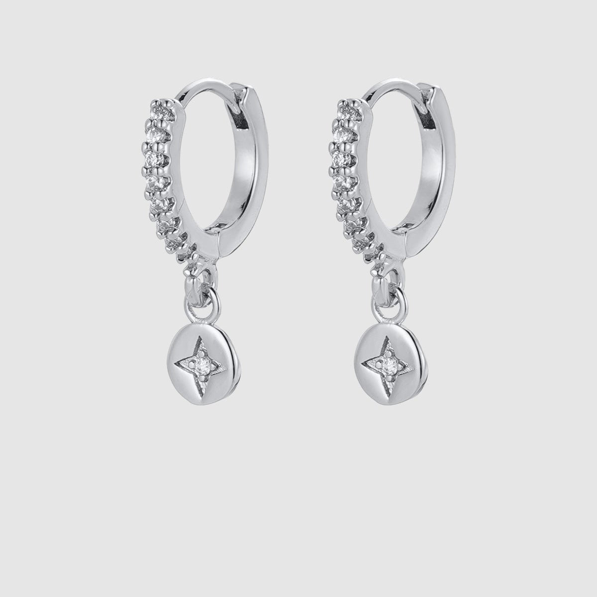 A pair of silver huggie hoop earrings with a curved row of small clear stones and a dangling round star-shaped charm, shown on a light gray background.