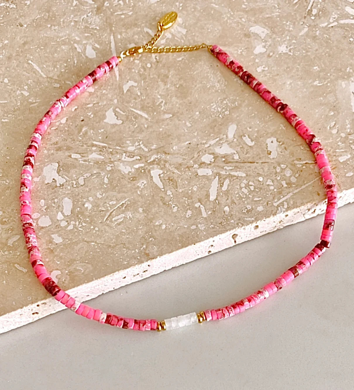 A beaded necklace made of small pink and red faceted beads with gold spacer rings and a gold clasp, displayed on a light beige stone surface.