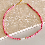 A beaded necklace made of small pink and red faceted beads with gold spacer rings and a gold clasp, displayed on a light beige stone surface.