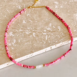 A beaded necklace made of small pink and red faceted beads with gold spacer rings and a gold clasp, displayed on a light beige stone surface.