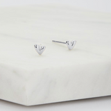 A pair of small polished silver heart stud earrings resting on a white marble block against a clean white background.