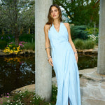 Woman in a light blue dress standing by a stone column with a garden and pond in the background
