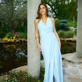 Woman in a light blue dress standing by a stone column with a garden and pond in the background