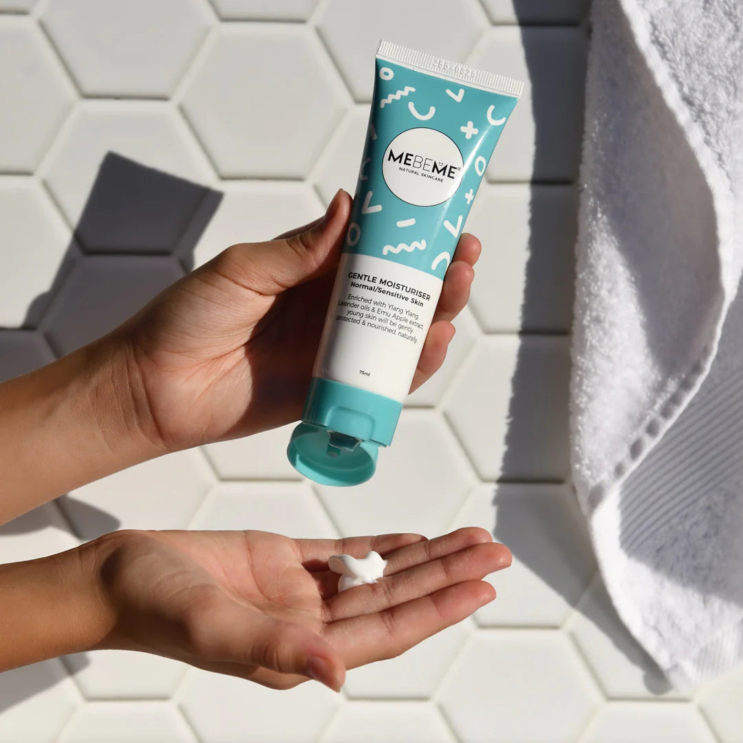 A person holds a tube of MEBEEME Gentle Moisturizer in one hand while squeezing a dollop of white cream into the other hand, set against a background of white hexagonal tiles and a white towel.